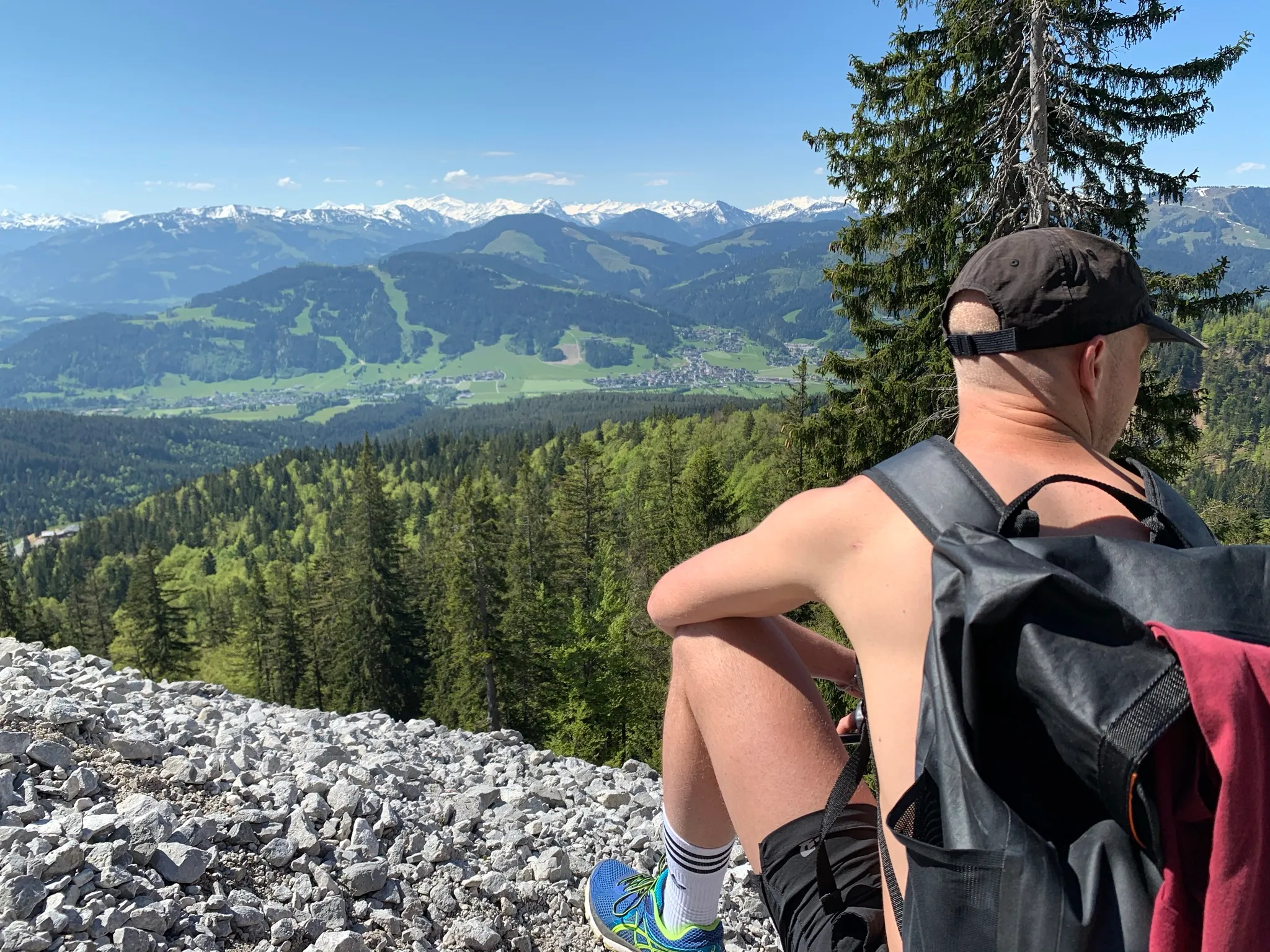 Ein IDALABS-Mitarbeiter sitzt auf dem Gipfel eines Berges und schaut in ein grün bewaldetes Tal hinab. Er trägt eine schwarze Cappy und einen Wanderrucksack. Im Hintergrund sind mit Schnee bedeckte Berggipfel zu sehen.