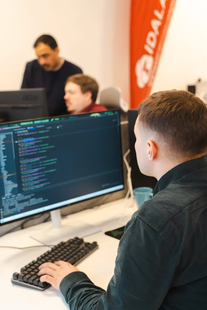 Ein junger Mann ist von hinten zu sehen, wie er Codezeilen an einem Computerbildschirm bearbeitet. Im Hintergrund sind weitere IDALABS-Mitarbeiter bei der Arbeit sowie ein rotes IDALABS-Banner zu erkennen.