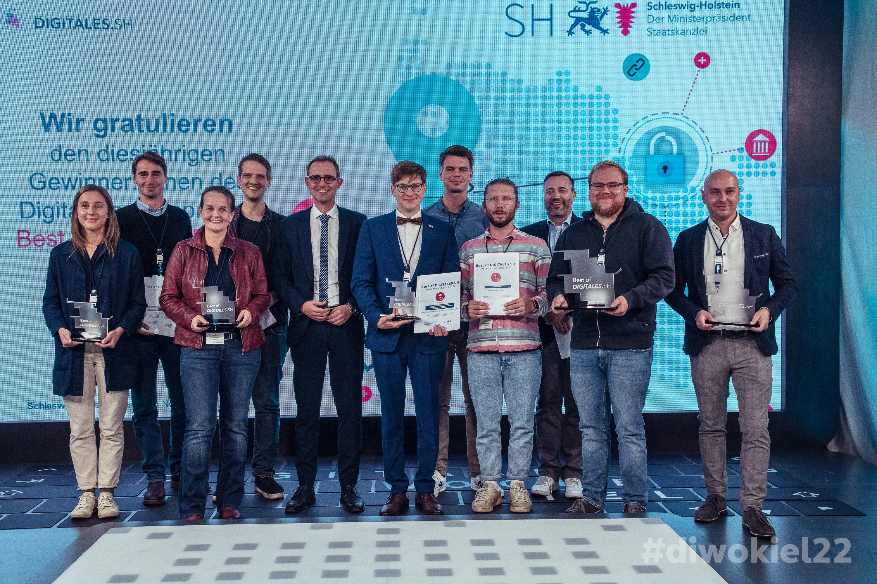Vor einer beleuchteten Leinwand stehen 11 Männer und Frauen, darunter die IDALABS-Gründer Alexander Westen und Tobias Gürtler. Sie zeigen stolz ihre Trophäen für die Auszeichnung zur “Best of Digitales S.H.”.