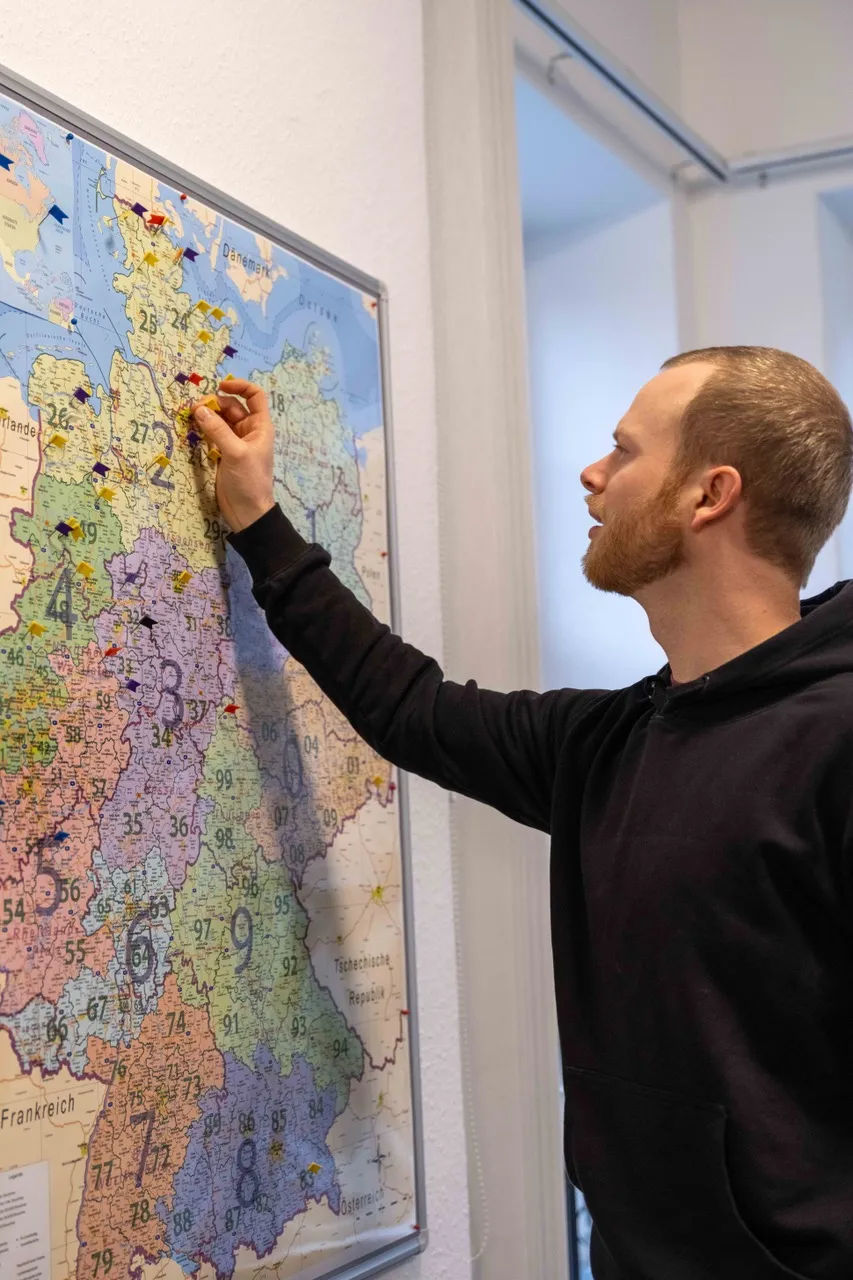 Ein Mitarbeiter der IDALABS steckt eine Reiszwecke mit einem kleinen Fähnchen in eine Deutschlandkarte. Die Fähnchen zeigen die Standorte unserer Kundinnen und Kunden an.