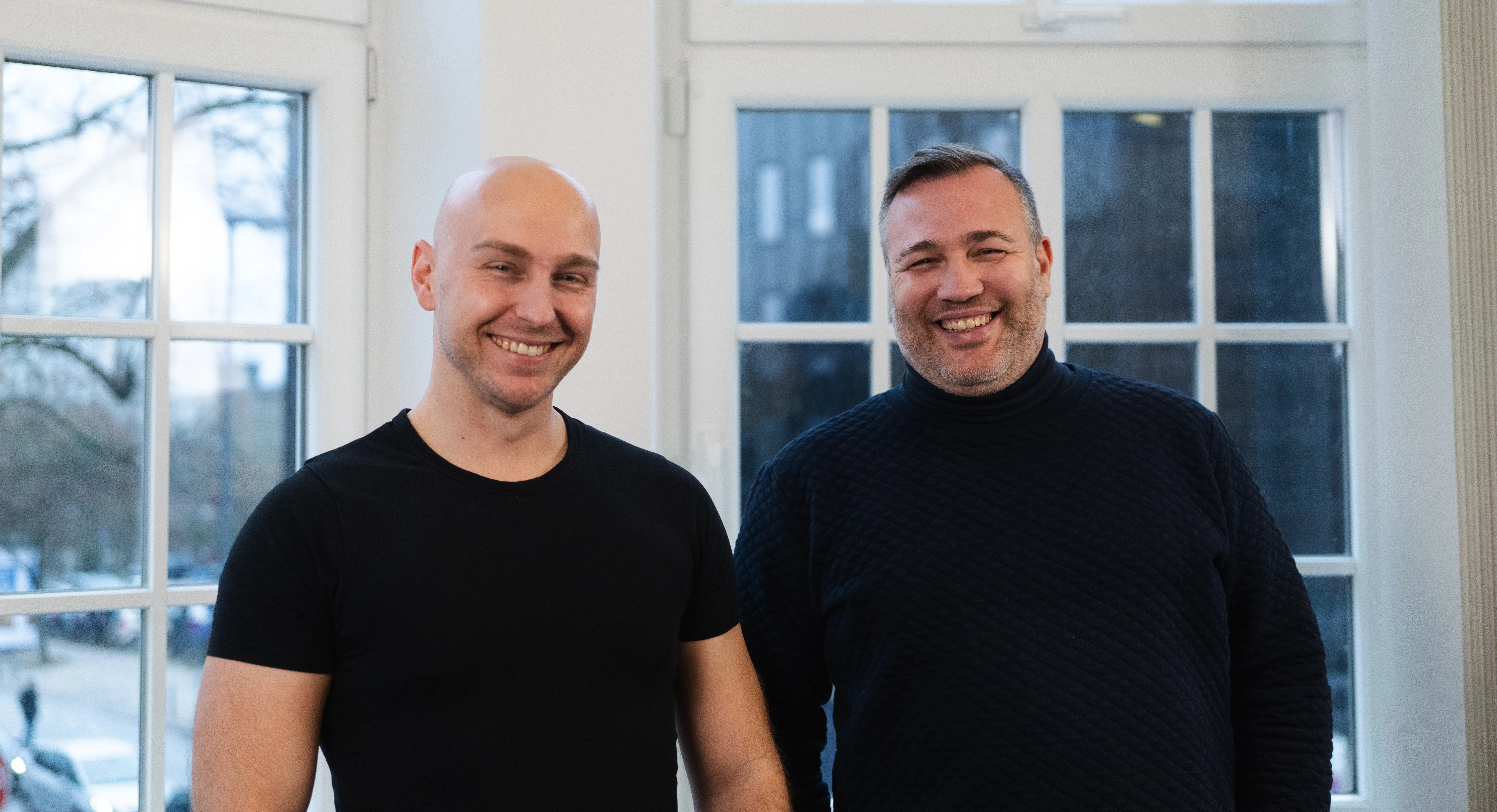 IDALABS-Gründer Alexander Westen und Tobias Gürtler stehen vor einer Fensterfront im Kieler IDALABS-Büro und lachen breit in die Kamera. Alexander Westen trägt eine Glatze, einen Dreitagebart und ein schwarzes T-Shirt. Tobias trägt kurze, grau-melierte Haare, einen Dreitagebart und einen blauen Rollkragenpullover.