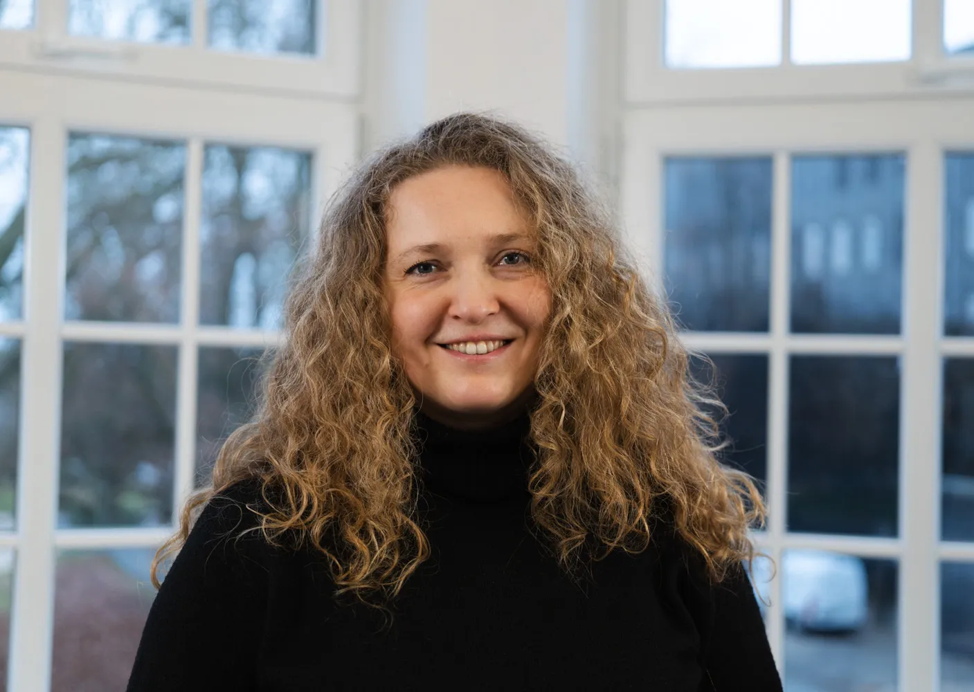 Patrycja Kupic lacht in die Kamera. Sie hat dunkelblonde Locken und trägt einen schwarzen Rollkragenpullover.