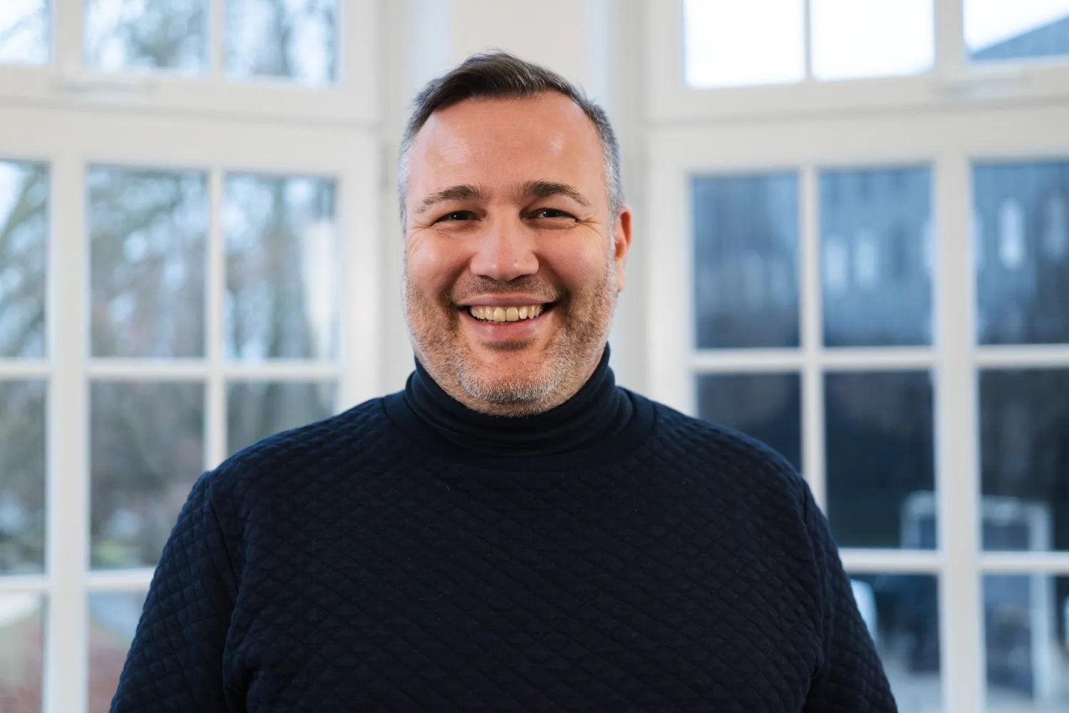 IDALABS-Gründer Tobias Gürtler lacht breit in die Kamera. Tobias trägt kurze, grau-melierte Haare, einen Dreitagebart und einen blauen Rollkragenpullover.