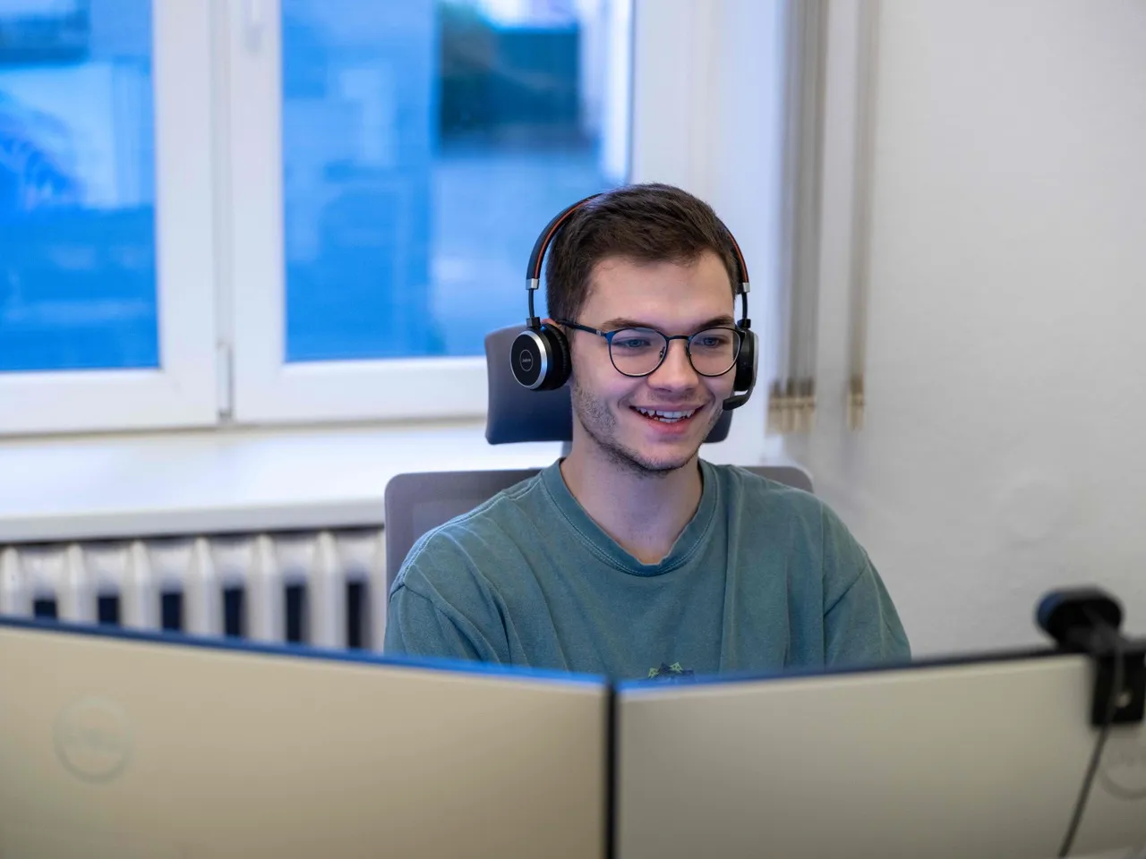 Lennart Leopold sitzt im grünen T-shirt vor zwei Computerbildschirmen in einem Büro. Er trägt ein Headset mit Mikrofon und lacht fröhlich.
