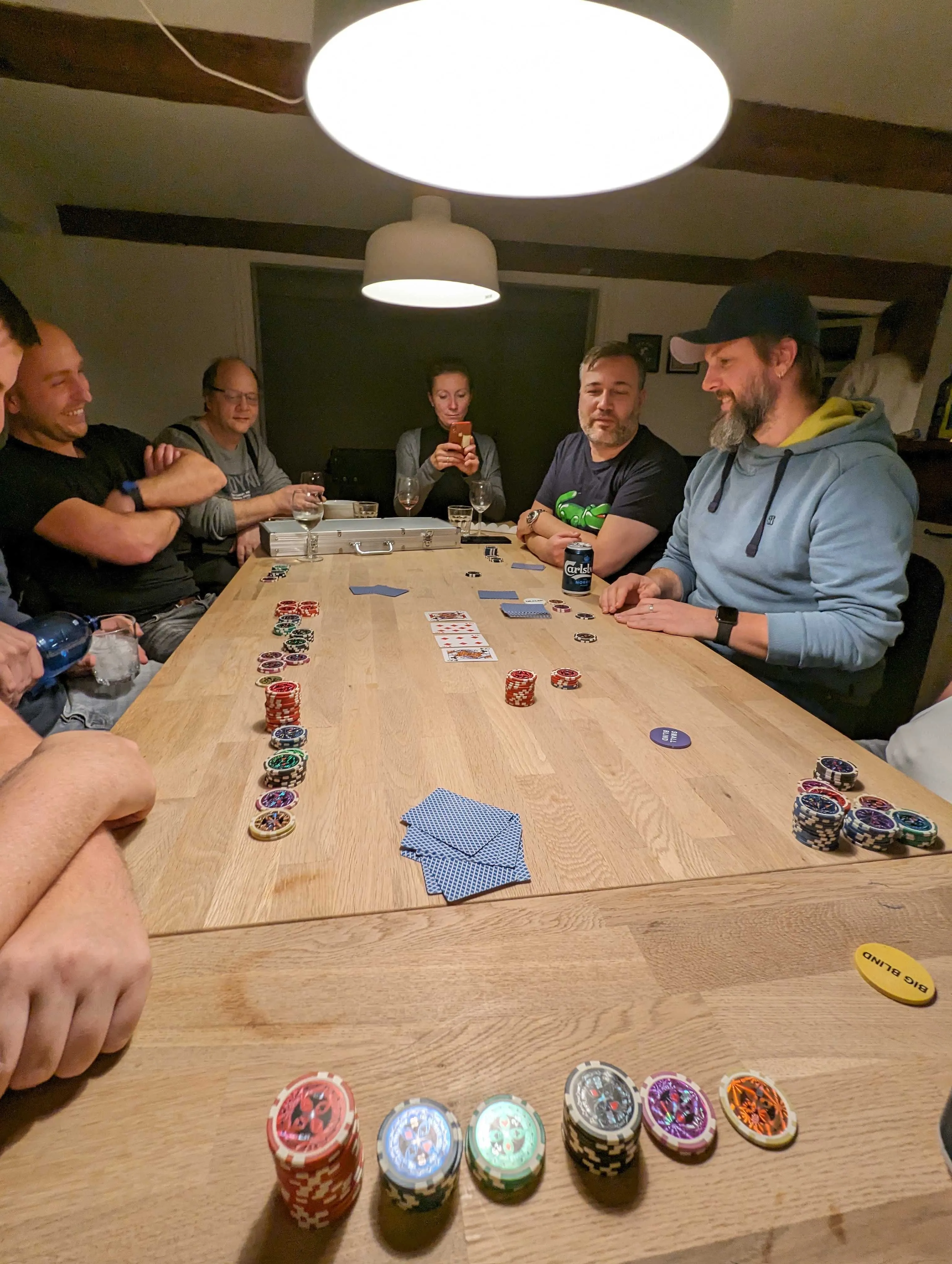  Fünf Männer und eine Frau sitzen an einem langen Holztisch. Vor ihnen sind verschiedenfarbige Poker-Chips gestapelt. In der Mitte sind vier Spielkarten aufgedeckt.