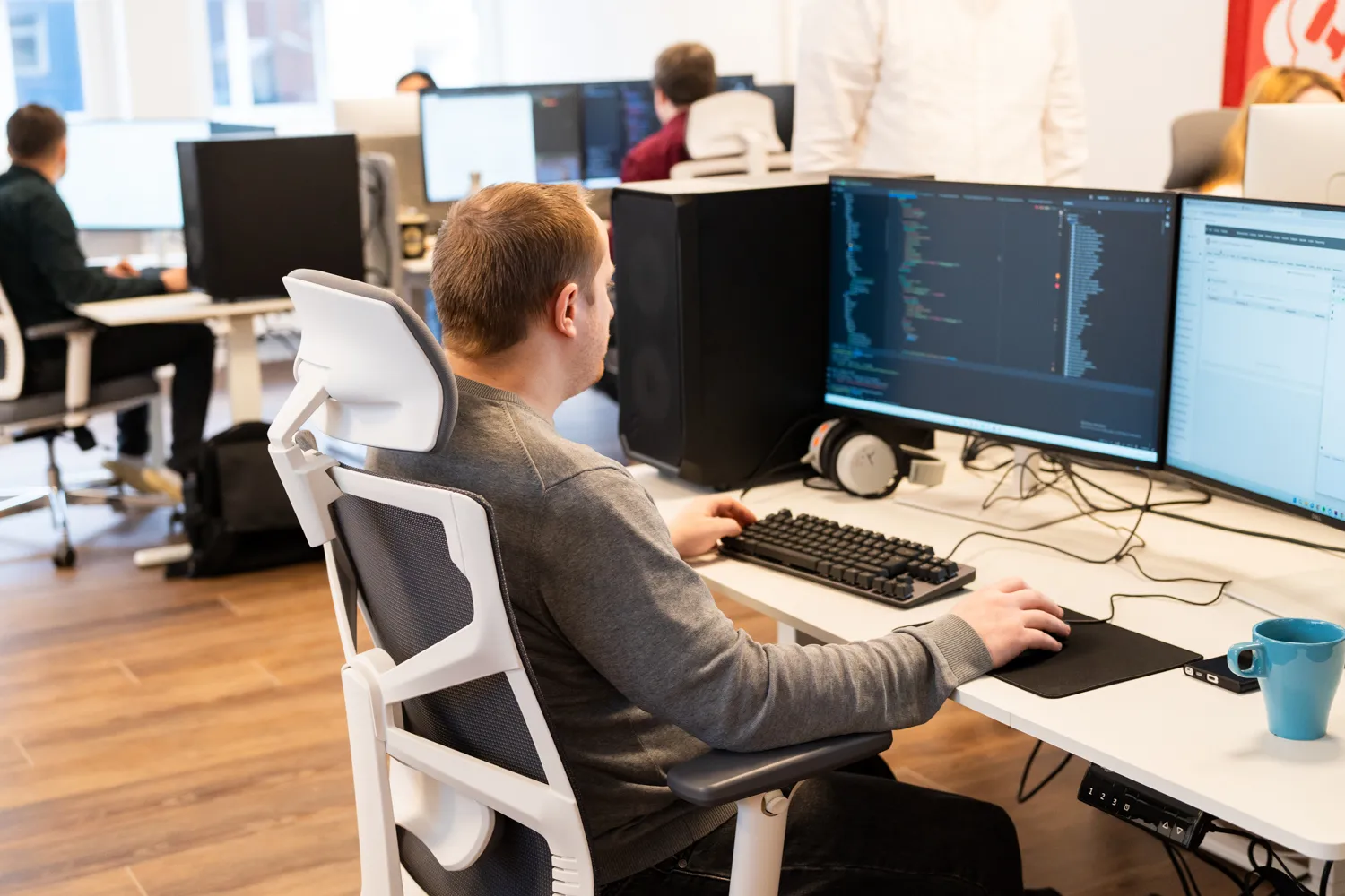 In einem hellen Großraumbüro sind sechs Mitarbeiterinnen und Mitarbeiter der IDALABS zu sehen, die auf ergonomischen Stühlen sitzen und teilweise an mehreren Bildschirmen gleichzeitig arbeiten.