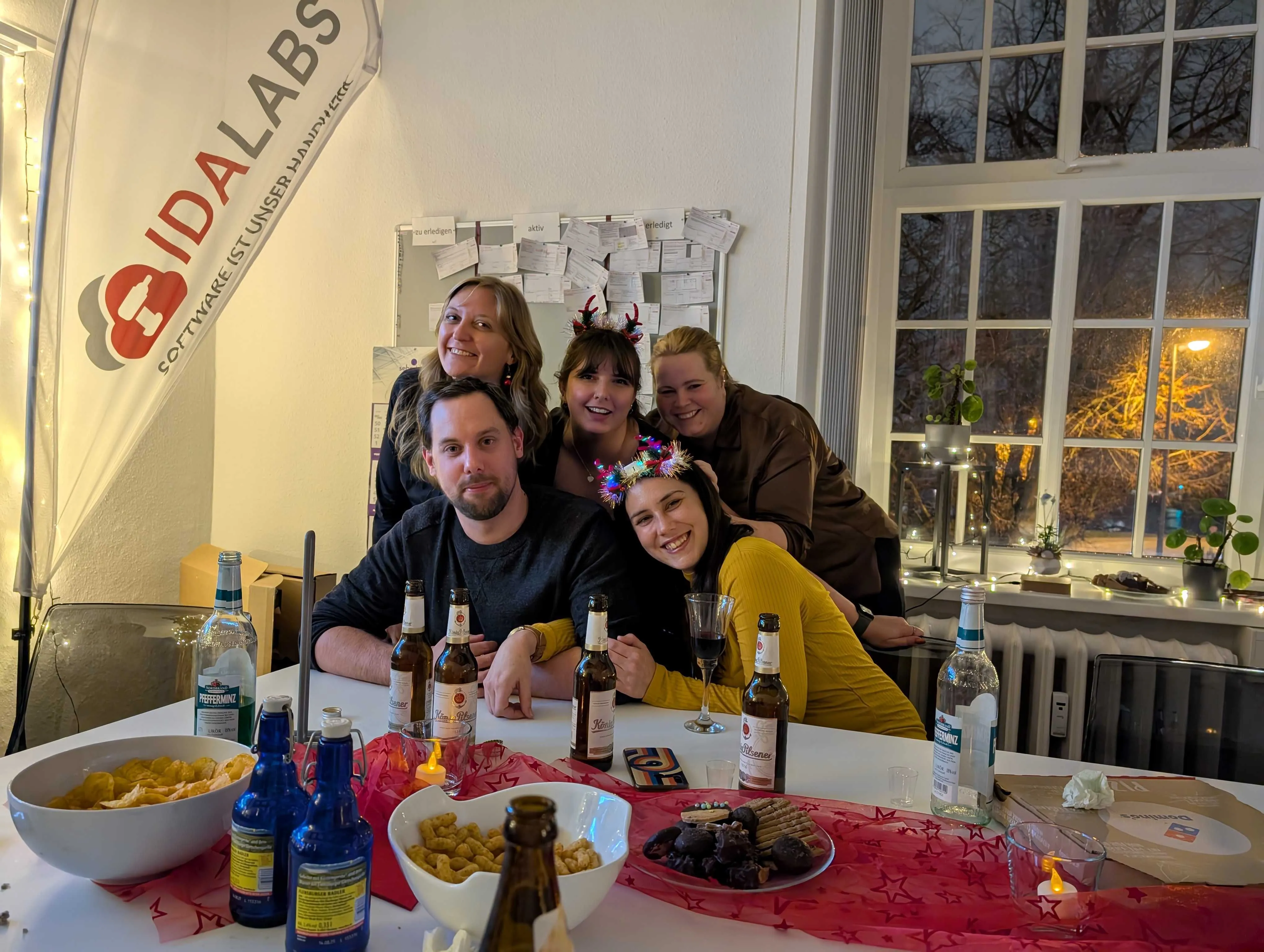  Vier junge Frauen und ein junger Mann kuscheln sich hinter einem weißen Konferenztisch zusammen und grinsen in die Kamera. Sie tragen Kopfschmuck mit weihnachtlicher Dekoration. Auf dem Konferenztisch stehen Bierflaschen, Schüsseln mit Chips und Weihnachtsschokolade.